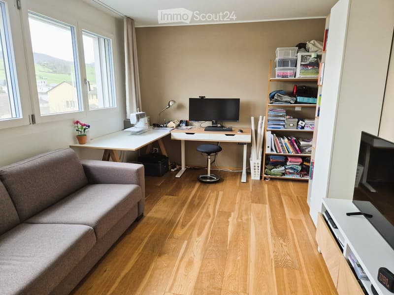 A well-lit room with wooden flooring, a grey sofa, a desk with a laptop, a stool, a shelf with books and folded clothes, and a flower pot on the desk.
