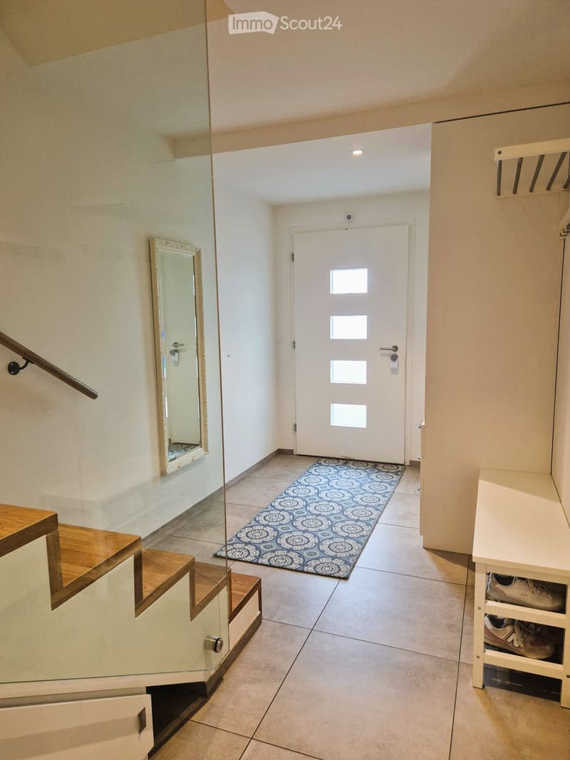 Modern hallway with tiled floor, glass staircase with wooden steps, white door with four windows, patterned rug, and shoe rack with shoes.
