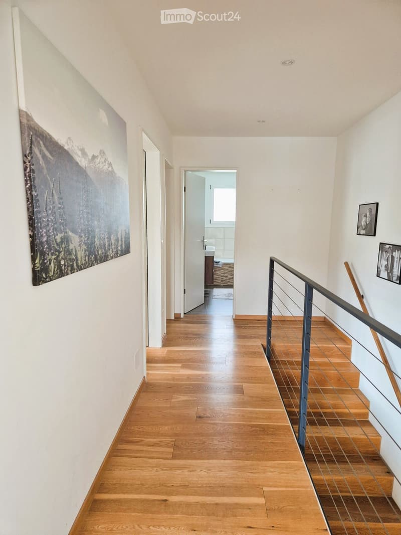 Spacious hallway with wooden flooring, white walls, and a modern staircase with metal railing leading upwards. A large painting and framed photos adorn the walls, and a partially visible bathroom is accessible from the hallway.