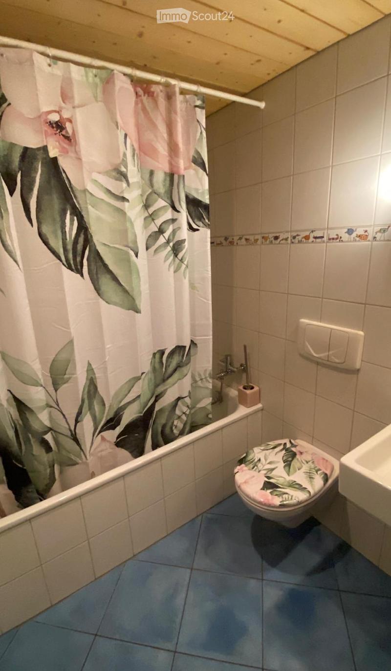Bathroom with a bathtub, toilet, floral shower curtain, and a floral toilet seat cover. Blue tiled floor and beige tiled walls.