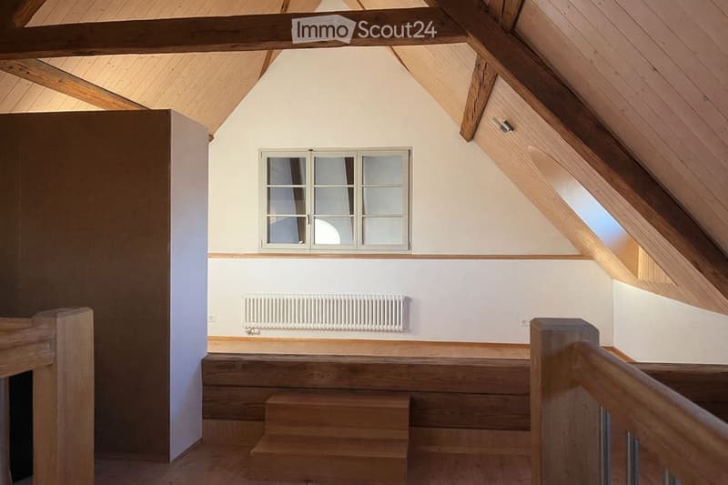 Empty attic room with wooden beams, exposed wooden ceiling, wooden staircase, white walls, and a window with frosted glass panels.