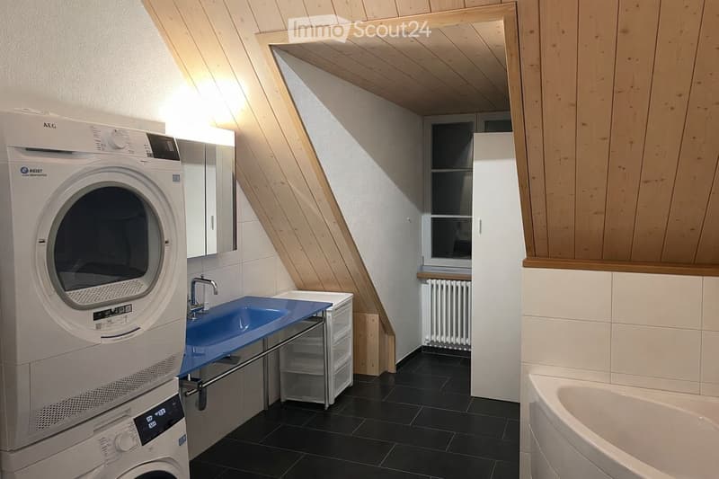 Modern bathroom with a washing machine and dryer, a blue sink, a mirror, a bathtub, and a radiator. The room has wooden slanted ceilings and tiled walls.