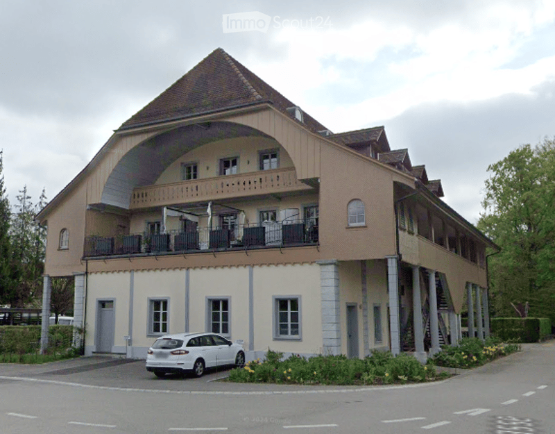 Two-story building with a curved facade, multiple windows, balconies, and a parked car in front.