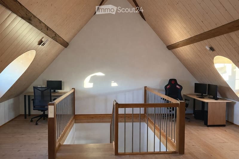 Bright attic room with wooden beams, slanted ceilings, and large windows. Contains two desks with computer monitors and chairs, separated by a wooden railing.