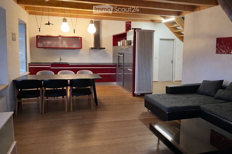 Open-plan living and dining area with wooden flooring, exposed wooden beams, a grey sectional sofa, a glass coffee table, a dining table with four chairs, and a modern kitchen with red cabinets and stainless steel appliances.