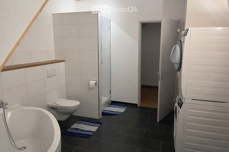 Modern bathroom with a bathtub, toilet, glass-enclosed shower, and a washing machine in the corner. The space features white tiled walls, dark tiled flooring, and two small rugs.