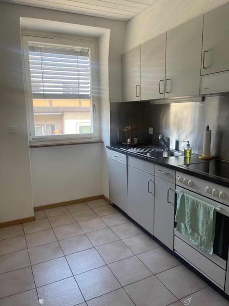 Modern kitchen with white cabinets, stainless steel appliances, tiled floor, and a window with blinds. Includes a sink, oven, dishwasher, and a towel hanging on the oven door.