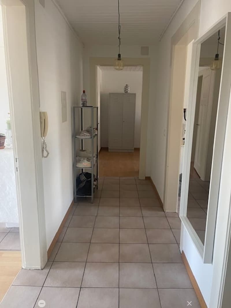 A clean, well-lit hallway with tiled flooring, white walls, and a small metal shelf holding shoes and a bottle. A closed door leads to another room.