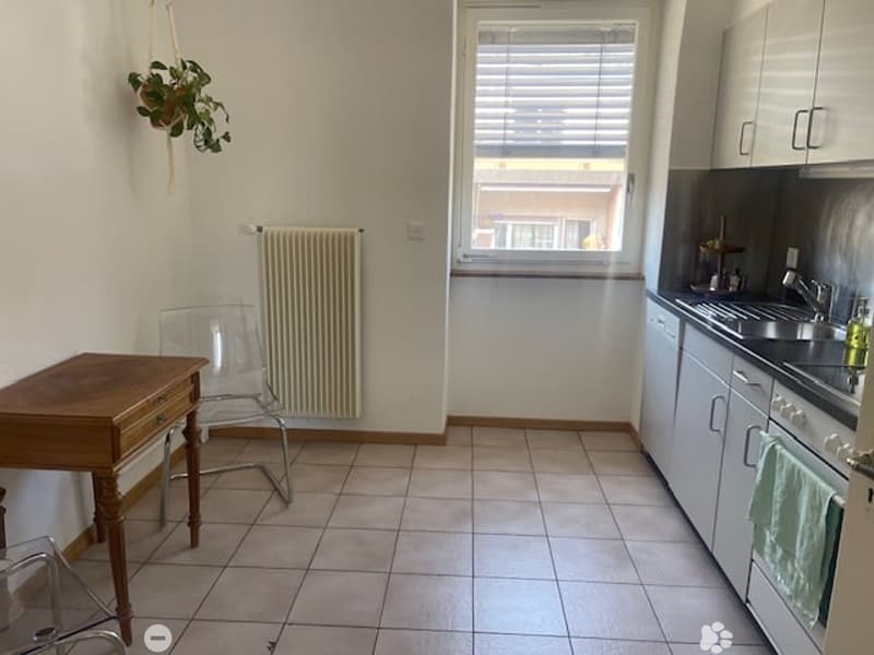 Bright kitchen with tiled floor, white cabinets, sink, oven, and a small wooden table with a chair. A radiator and a hanging plant are visible.