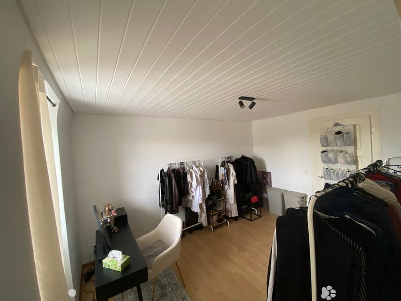 A well-lit room with wooden flooring, a white chair, a black table with a screen, and clothing racks with various garments.