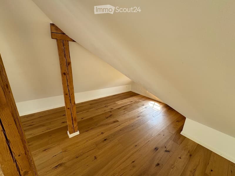 Empty attic space with wooden flooring and exposed wooden beams, featuring sloped ceilings and natural light from a small window.