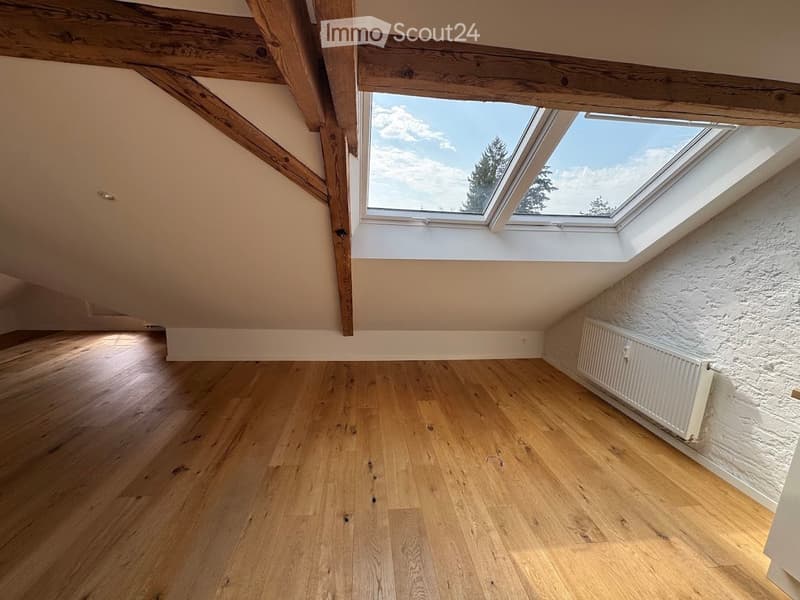 Empty attic room with wooden flooring, exposed wooden beams, and skylight windows offering a view of the sky and trees.