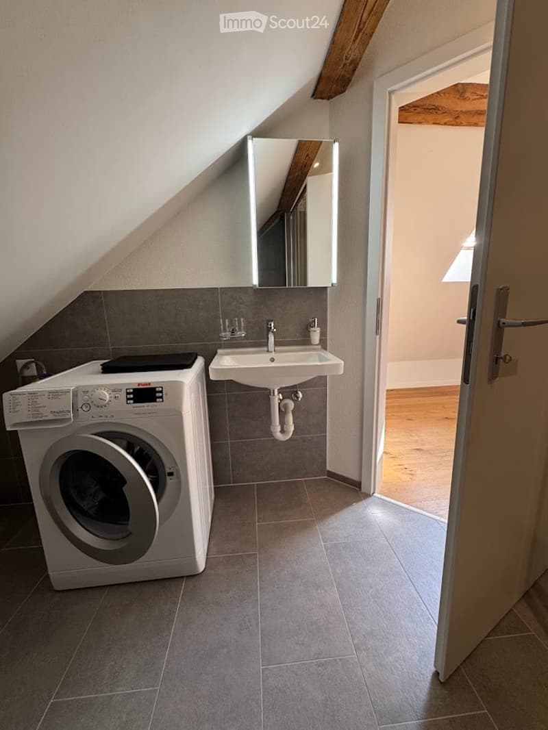 Modern bathroom with a washing machine, sink, and mirror. The room features grey tiled walls and floor, with a slanted ceiling and wooden beams.