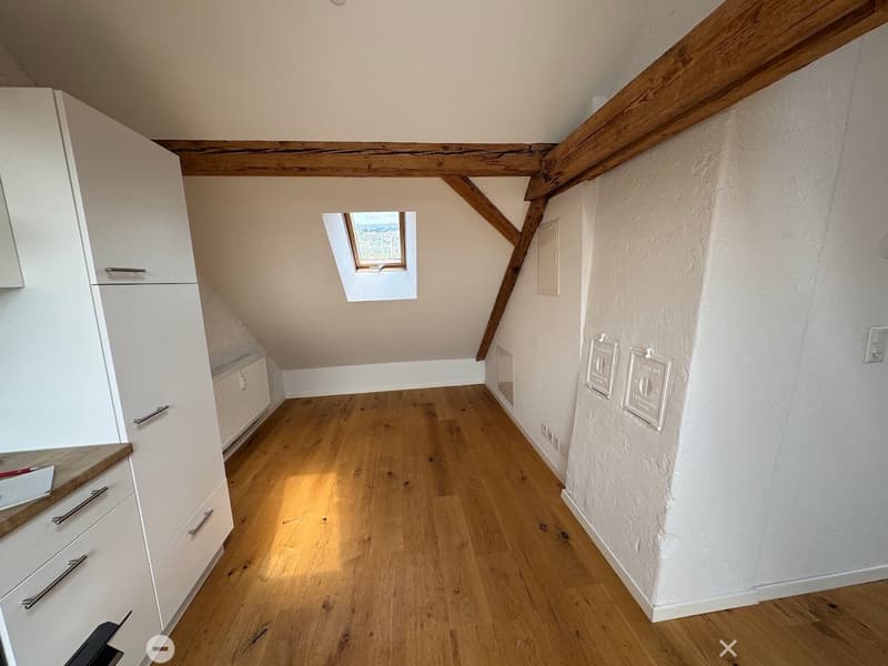 An unfurnished attic room with wooden floors, exposed wooden beams, and a skylight window. The room features white cabinetry on one side and a small staircase leading to another area.