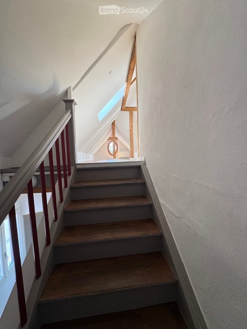 Wooden staircase with red handrail and white walls, leading to an attic space with skylight.
