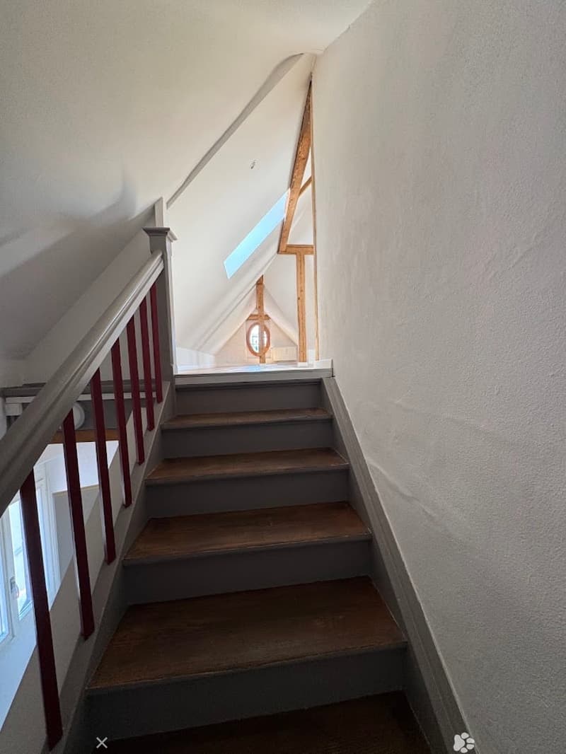 Wooden staircase with red handrail and white walls, leading to an attic space with skylight.