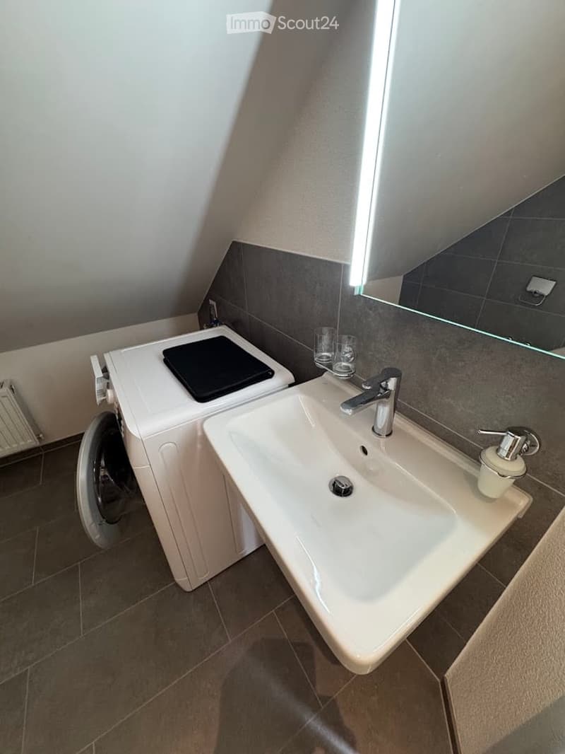 Modern bathroom with a white sink, chrome faucet, soap dispenser, and a washing machine with a black lid. The room features a large mirror, tiled walls, and a slanted ceiling typical of an attic space.