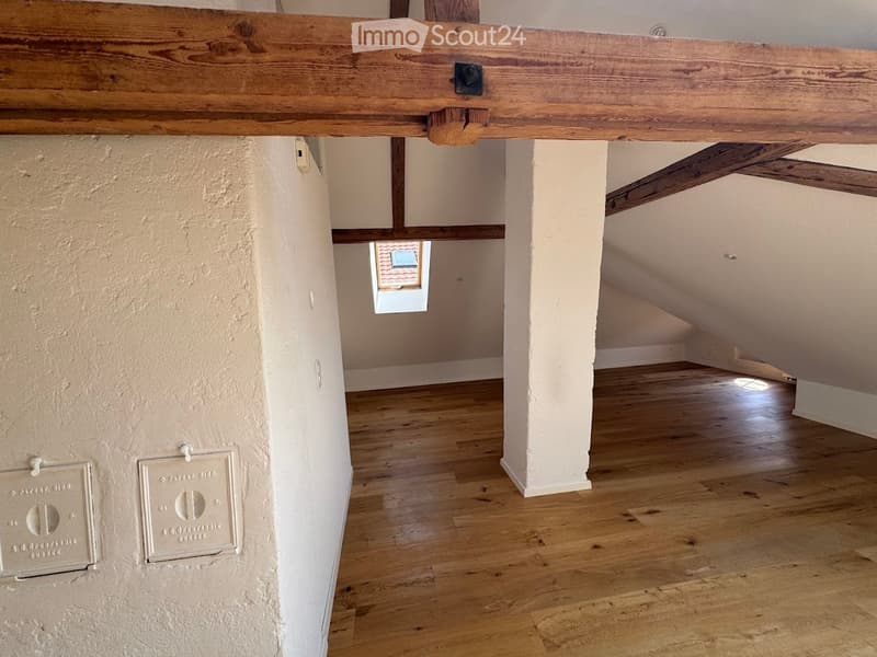 Empty attic space with wooden beams, exposed wooden floor, and a small window letting in natural light.