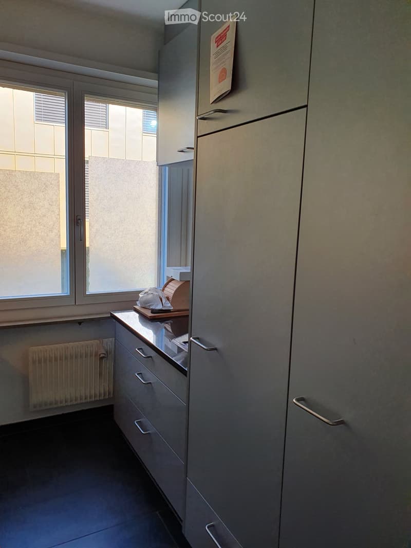 Modern kitchen with grey cabinets, countertop, and a small window. Includes a radiator and a few items on the counter.