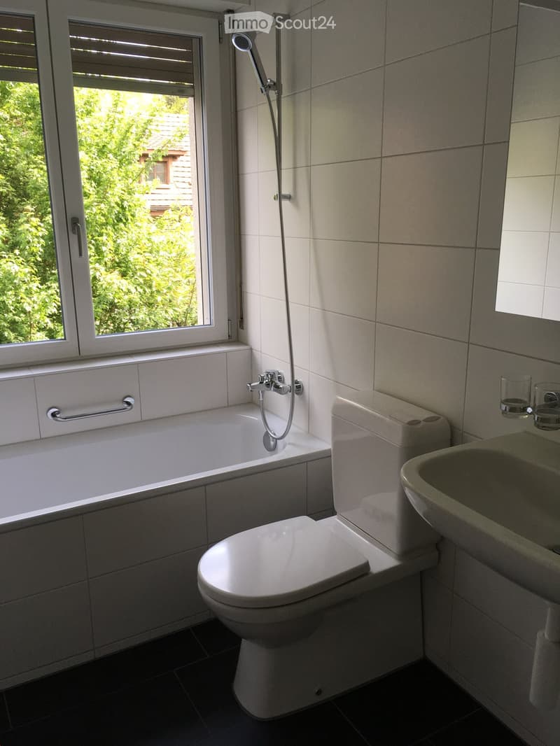 Modern bathroom with a bathtub, toilet, sink, and showerhead. The room features white tiles and a window with blinds, allowing natural light to enter.