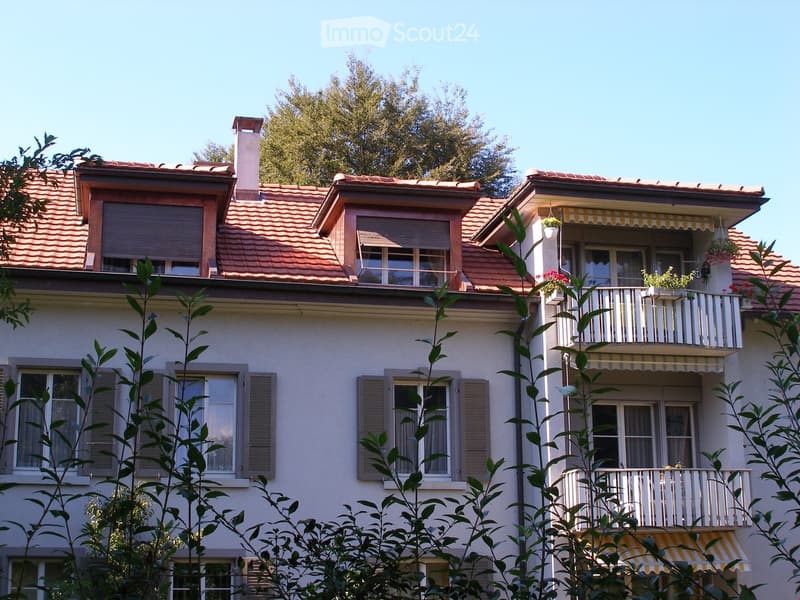 red tiled roof, white walls, wooden balcony with awning, multiple windows, greenery surrounding