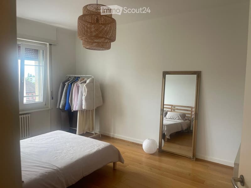 Bedroom with wooden flooring, a large mirror, a bed with white bedding, a wicker hanging lamp, a clothing rack with hanging clothes, and a small white spherical lamp on the floor.