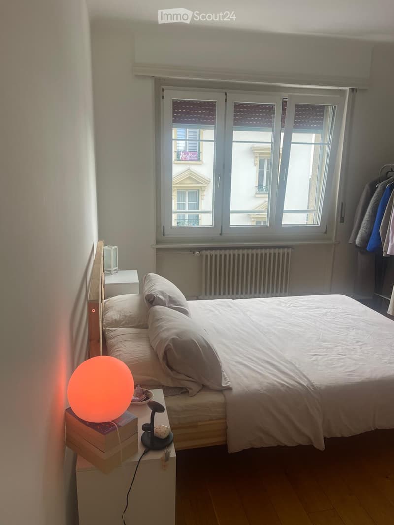 Cozy bedroom with a neatly made bed, nightstand with a lamp, and a round orange lamp on a stack of books. The room features wooden flooring and large windows with blinds.