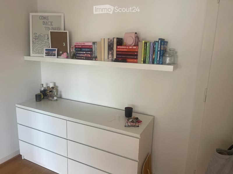 A neatly organized bedroom with a white dresser, a floating shelf holding books and decorative items, and a small table with perfume bottles and a cup.