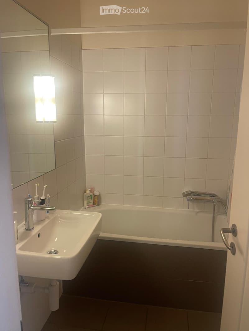 Modern bathroom with white tiles, a sink with a chrome faucet, a bathtub, a mirror, and personal care items on the sink counter.
