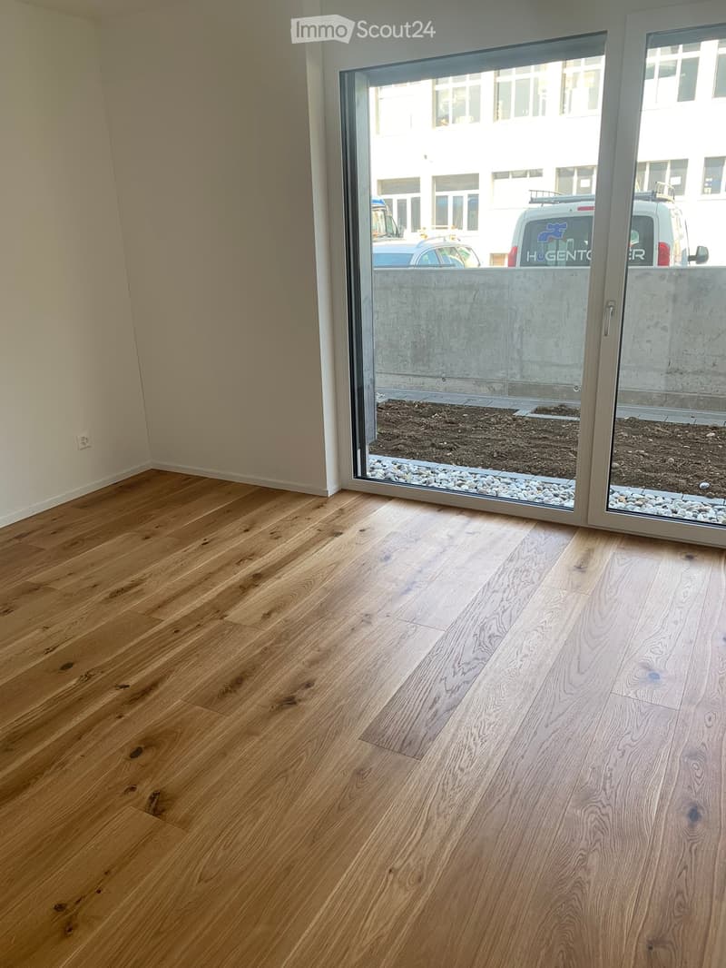 Empty room with light wooden flooring and a large glass door leading to an outdoor area with gravel and a concrete wall.