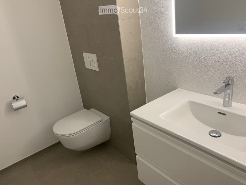 Modern bathroom with a white toilet, a white sink with a chrome faucet, and a window above the sink. The walls are tiled in neutral tones, and the floor is tiled in a light brown color.