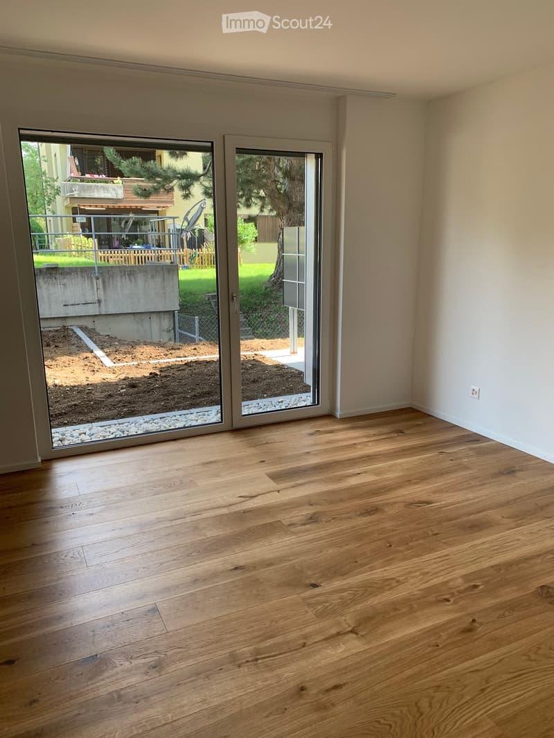 Empty room with wooden flooring and large sliding glass doors leading to an outdoor area with gravel and a concrete structure.