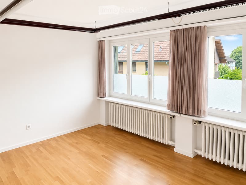 Empty room with wooden flooring, white walls, large windows with brown curtains, and radiators beneath the windows.