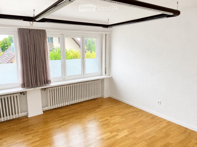 Empty room with wooden flooring, white walls, large windows with grey curtains, radiator, and exposed ceiling beams.