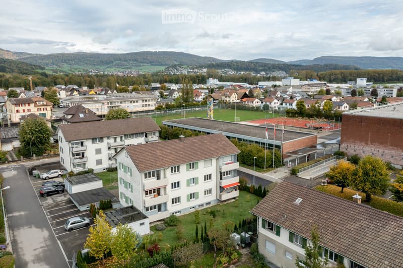 Aerial view of a residential area with multiple apartment buildings, parking lots, and surrounding greenery. The buildings have brown tiled roofs and white facades, with some featuring balconies. The area includes a school complex, sports fields, and a di