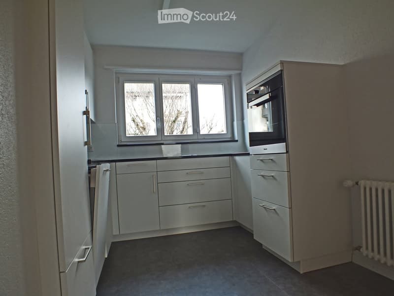 Modern kitchen with white cabinets, a built-in oven, and a radiator. The space features a large window with three panes and a dark tiled floor.