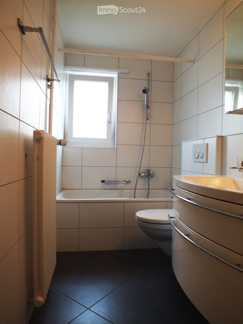 Modern bathroom with white tiles, dark tiled floor, bathtub, toilet, sink, and a window above the bathtub.