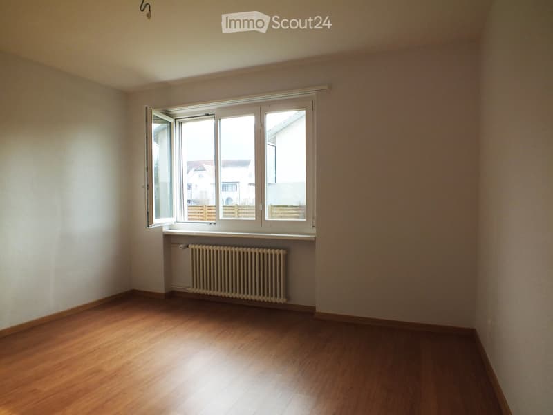 Empty room with wooden flooring, white walls, and a large window with white frames. A radiator is visible below the window.