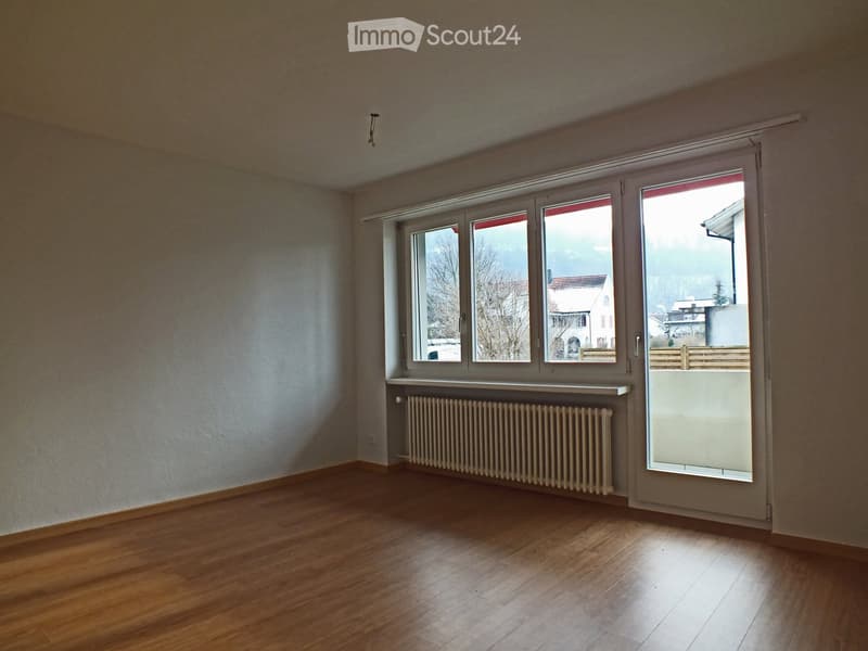 Empty room with wooden flooring, white walls, large windows, radiator, and a sliding door leading to a balcony.