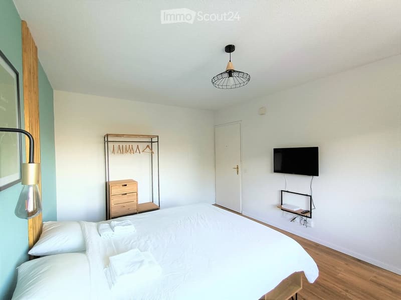 Cozy bedroom with a neatly made double bed, white pillows, folded towels, a wooden dresser with a hanger rack, a wall-mounted TV, and modern lighting fixtures.