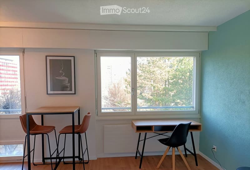 A small, well-lit room with light blue and beige walls, wooden flooring, and large windows offering a view of trees. Contains a small table with two chairs, a stool, and a picture frame on the wall.