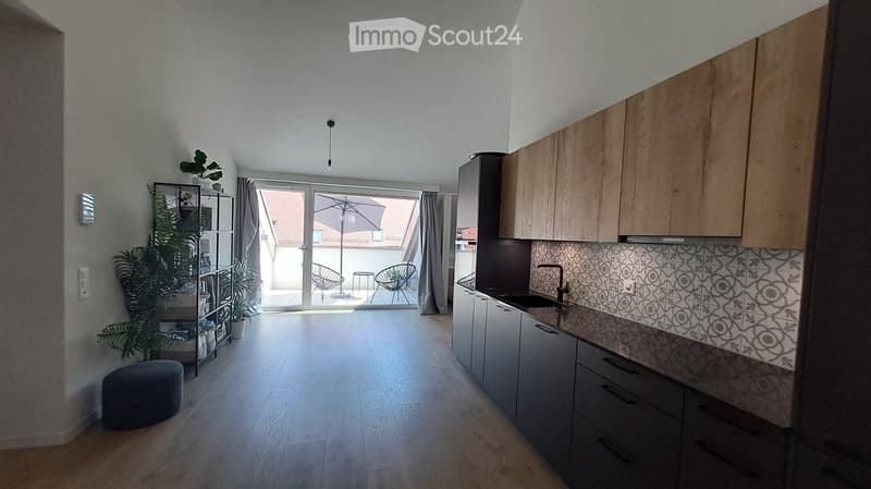 Modern kitchen with wooden cabinets, stainless steel sink, and a small seating area with a stool and plants near the sliding glass door leading to a balcony.