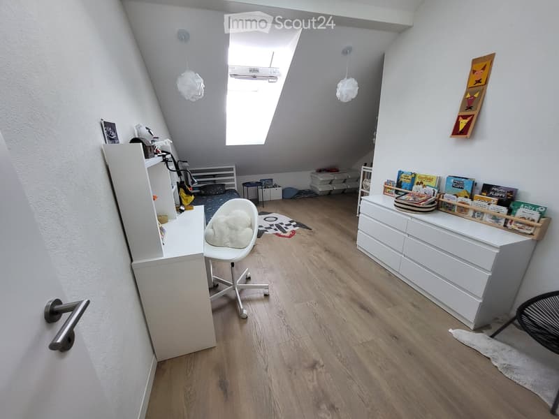 Bright, modern attic bedroom with wooden flooring, slanted ceilings, and large skylight. Features a white desk, bookshelf, bed, and storage unit.