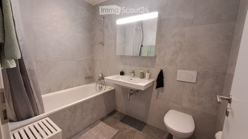 Modern bathroom with a bathtub, sink, toilet, and a large mirror with built-in lighting. The walls are tiled in a light gray color, and there are towels and small decorative items on the sink counter.