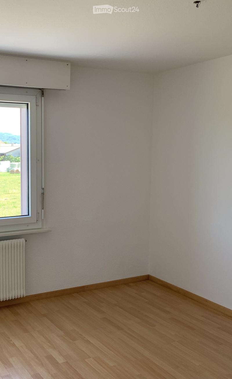Empty room with wooden flooring, white walls, a window with a view of the outdoors, and a radiator below the window.