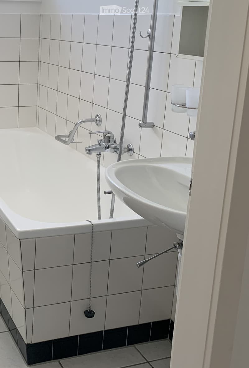 Modern bathroom with a white bathtub, a sink, and a shower hose attached to the wall. The tiles are white with a black baseboard. A cup and cup holder are visible on the right side.