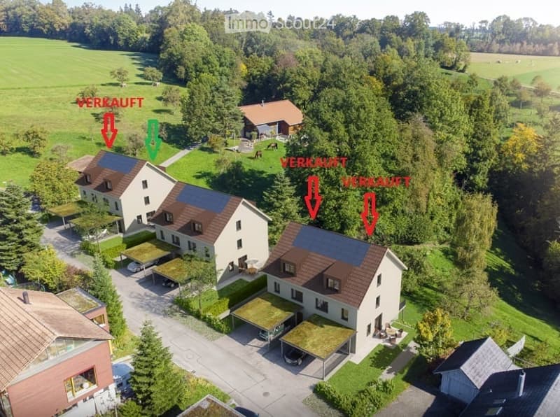 Aerial view of a residential area with multiple houses, green lawns, trees, and driveways. Some houses have solar panels on the roofs.