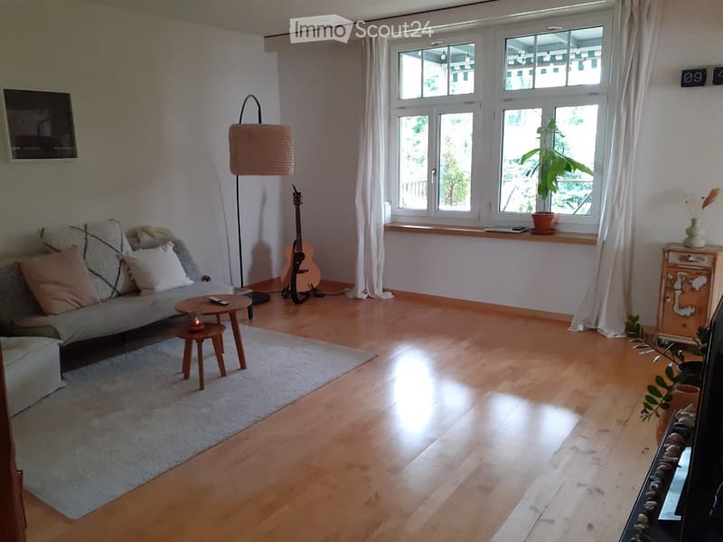 Living room with wooden flooring, large windows, sofa, coffee table, guitar, floor lamp, and decorative plants.