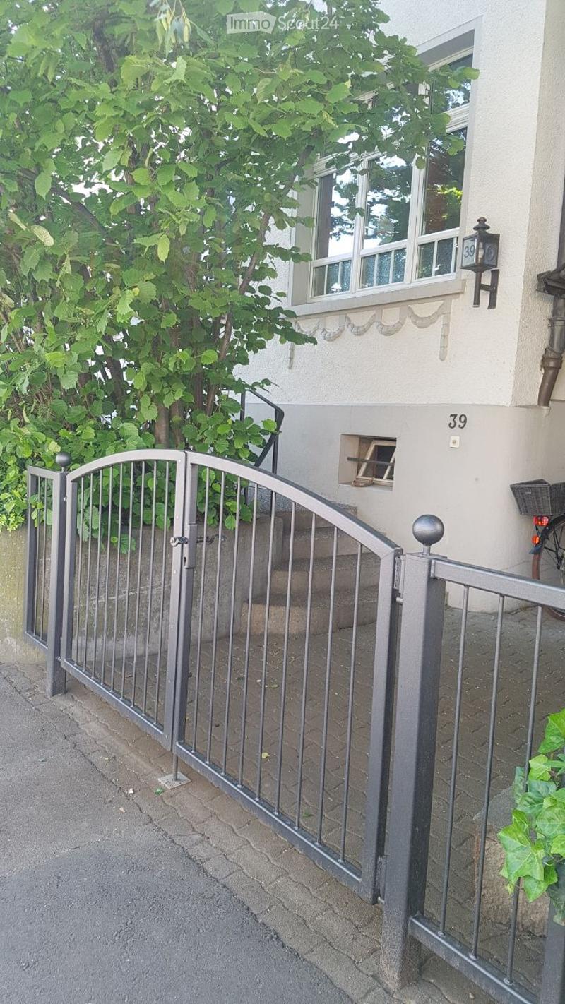 A raised entrance gate leads up to a house with a staircase, surrounded by greenery and a tree.