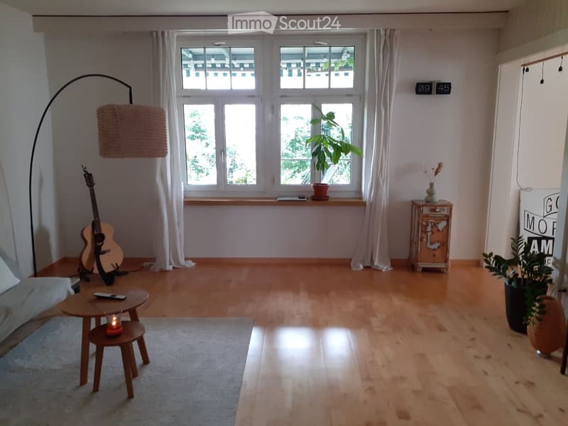 Living room with wooden floor, large windows, sofa, guitar, small table, floor lamp, and plants.